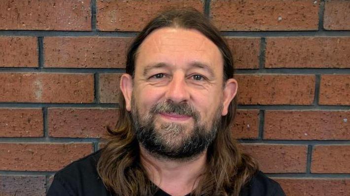 A head and shoulders picture of Lou Chawner. He has long brown hair and a short black and grey beard. He is wearing a black T-shirt and is standing in front of a red bricked wall.