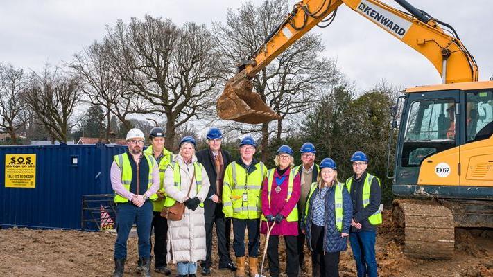 Nine people are stood in a line at a construction site, all wearing hi vis jackets and construction hats. Behind them is a large dark blue shipping crate with a yellow sign reading "S.O.S" in big black letters. There is also a yellow crane in the right side in the background. In the further background are many trees against a grey sky, most of them without leaves.