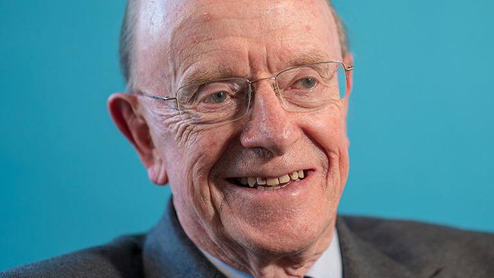 A smiling older man with glasses and a receding hairline against a blue background