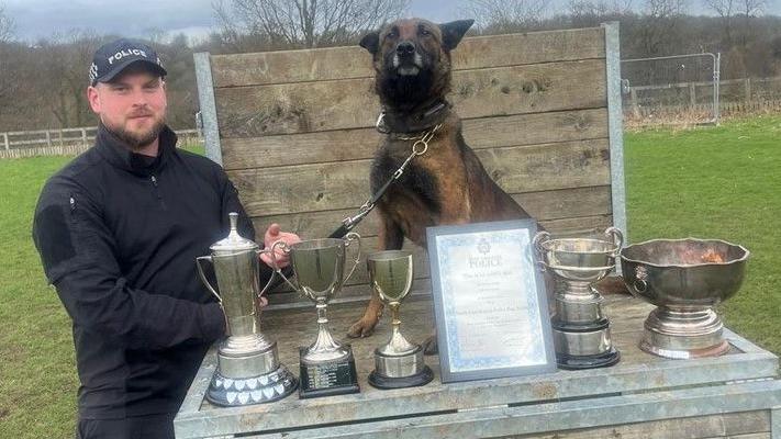 Quinta sits on a platform so is at the same level as her handler. There are five trophies and a certificate in front of her. She holds her head up high with her ears held back. PC Lusby holds her lead and stands on the left of the picture. He is dressed in black and wears a police cap.