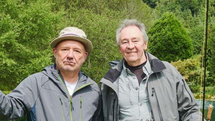 Paul and Bob standing next to each other looking towards the camera. They are both holding a fishing rod and wearing a greyish coat. Both surrounded by bright green trees, white daises and a dark green river behind them.