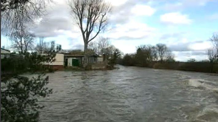 Mobile homes to the left of the picture, surrounded by fast-flowing water. There are trees in the background. There is visible wash in the water to the right.