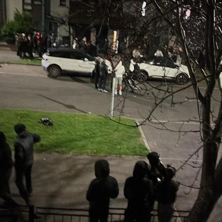 A shot of Hay Avenue showing a group of about 20 youths, all with their hoods up turned away from the camera. A number are standing around two white cars parked on the street.