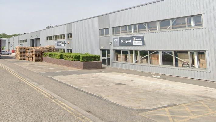 A view of three two-storey metal-clad industrial units with a road in front of them. There is a blue sky above.