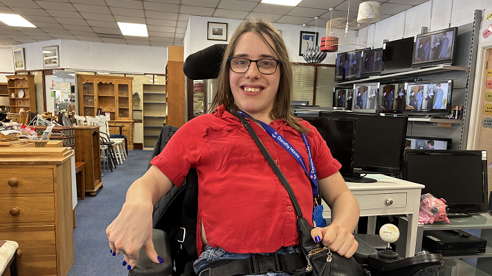 Freya Viles wears glasses and a red blouse and wears a blue Dorothy House lanyard while smiling at the camera. She is sat in a wheelchair inside a charity shop, with pieces of furniture and TVs around her. 