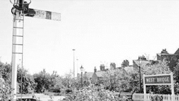 A black and white image of the West Bridge station platform with a sigal post in the foreground.