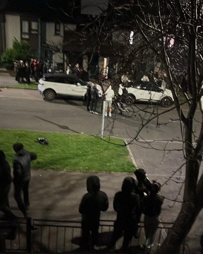 A shot of Hay Avenue showing a group of about 20 youths, all with their hoods up turned away from the camera. A number are standing around two white cars parked on the street.