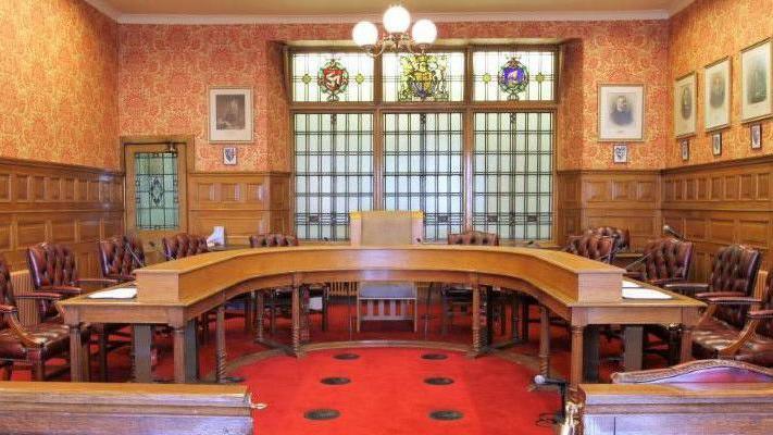 The Legislative Council chamber, which features a horseshoe-shaped wooden table and brown leather upholstered chairs with wooden arm rests. The room had a red carpet with two lines of three black spots down the middle. The walls are half wooden panelled with orange and yellow patterned wallpaper on the upper half. The are framed portraits on the right hand wall and stained glass windows at the far end of the room.