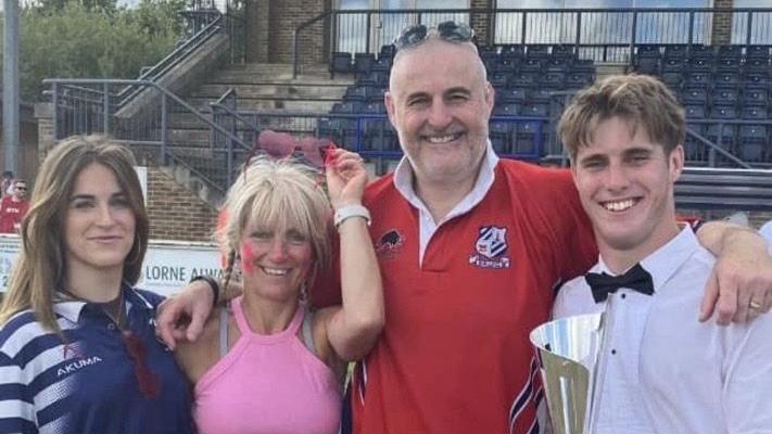 A woman with dark hair wearing a rugby shirt, a woman with blonde hair and wearing sunglasses on her head, a short-haired man wearing a red rugby shirt and a young man wearing a white shirt and a bow tie and holding a trophy, smiling at the camera with their arms around each other. They appear to be in a sports stadium with rows of seats behind them.