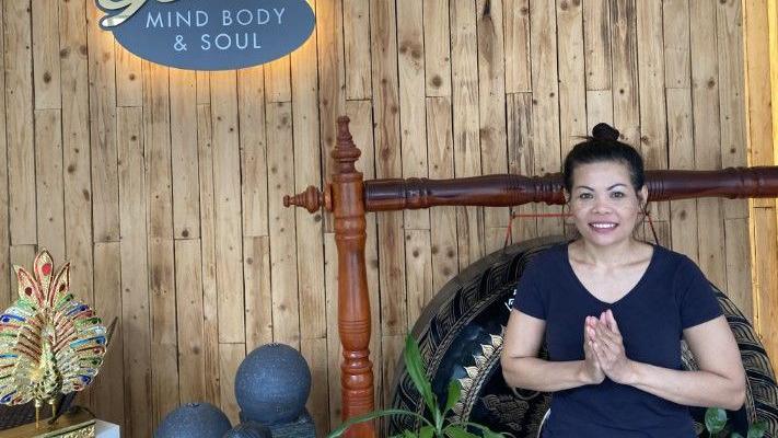 A woman in a black t-shirt, with her hair in a bun, presses her hands together upwards as she smiles at the camera. She stands in a room with wooden walls, with a sign saying 'mind, body & soul' on the wall