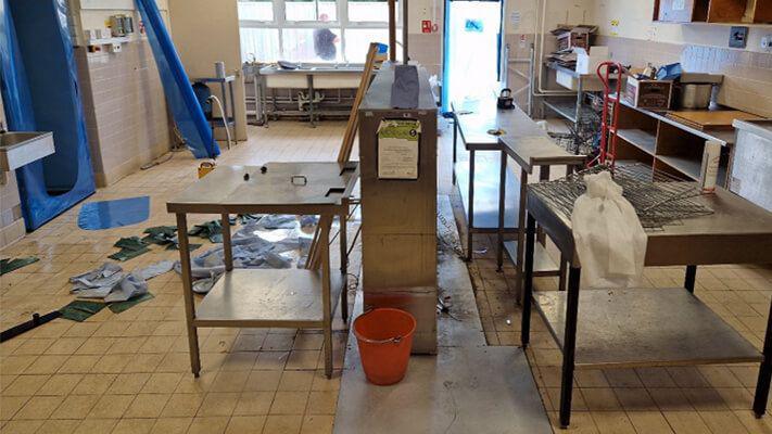 The inside of the school kitchen during construction. There are metal tables and grills in the centre of the tiled floor. The room is messy and there are paper towels all over the ground.