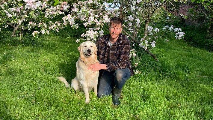 Liam is crouching down on the grass in the orchard alongside his dog Monty. They are both looking at the camera and Liam is smiling. Behind them is a tree which has blossomed with pale pink flowers.