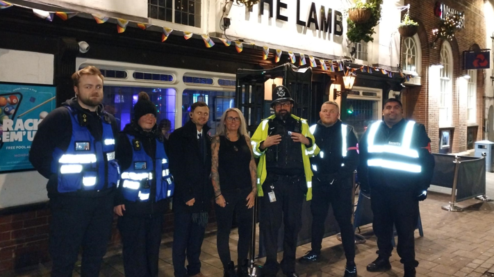 Seven people standing outside a pub at night, called the Lamb, one in hi viz jacket.