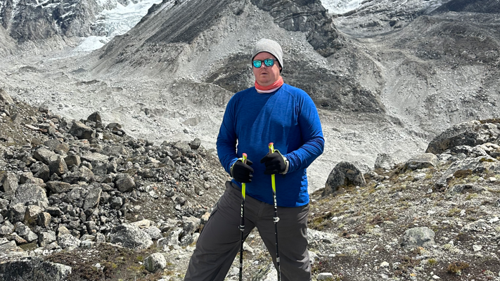 Simon Clark standing on rocks. He is wearing a blue shirt and a grey hat. He is leaning on walking sticks.