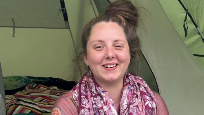 Emily Witts smiles while sat in the doorway of a green tent. She is wearing a maroon-coloured scarf with a pattern on it and a pink shirt with flowers on it. She has her hair in a beehive bun. A striped blanked is behind her in the tent.