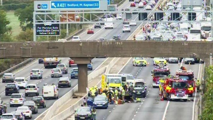 The M1 motorway, showing eight lanes of traffic, with vehicles on the road, and cars stopped. There are fire engines, ambulances and police vehicles on several lanes.