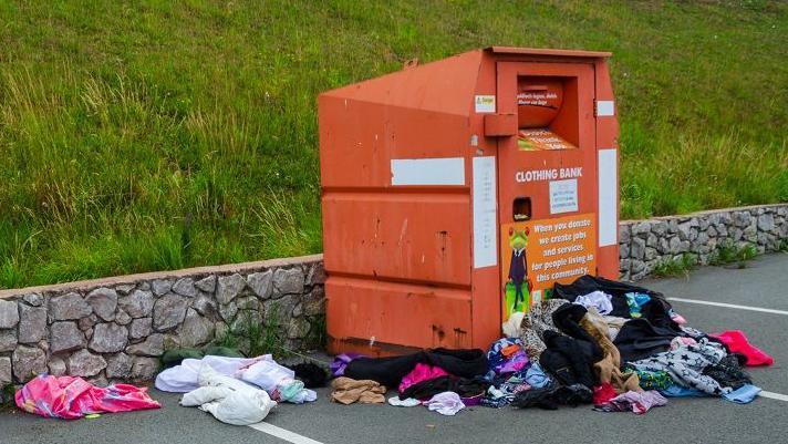 A large orange recycling container with piles of clothes dumped in front of and around it.