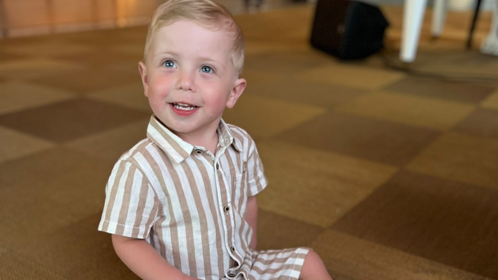 Hudson is wearing a brown and white stripy top and shorts and is sitting on the floor smiling at the camera
