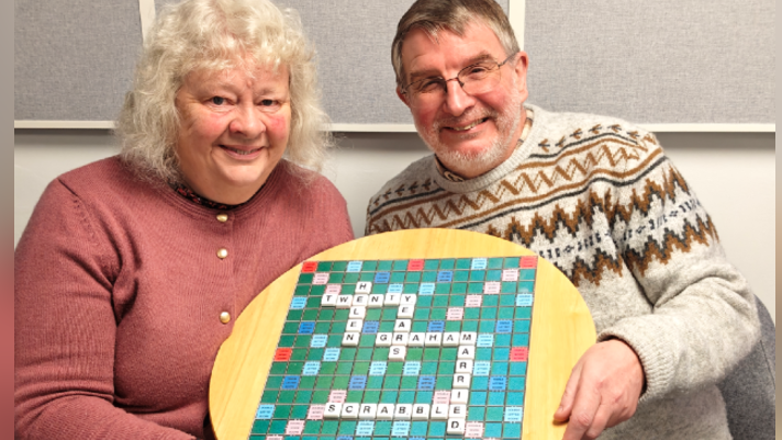 Helen is wearing a peach pink cardigan with golden buttons holding a scrabble board that has words Helen, Twenty Years, Graham, Married, Scrabble fitted in the scrabble board and Graham holding the Scrabble board as well and is wearing a crème coloured winter jumper. 