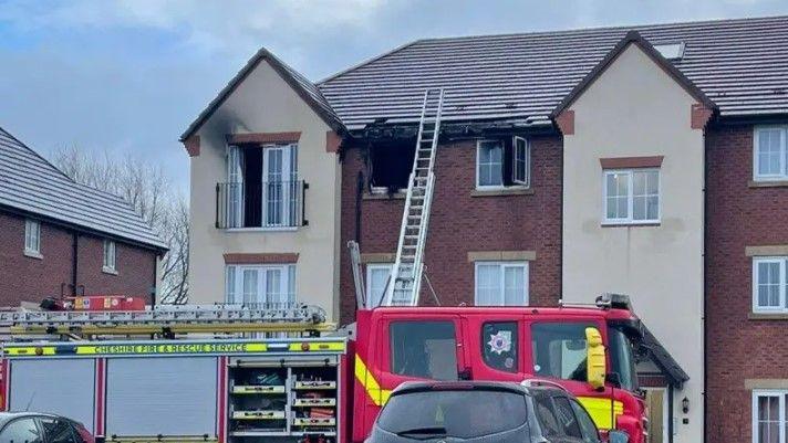 A fire engine in front of a flat with three burnt out windows. A ladder is leaning against the front of the property and reaches all the way up to the roof.