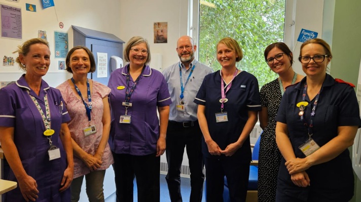 A photo which shows seven members of staff standing in a row smiling. The woman to the far left of the photo has a purple hospital uniform on, the woman next to her has a pink shirt on, the woman to the right of her has another purple uniform on. Next to her is a man wearing a blue shirt, and next to him is a woman wearing a blue hospital uniform. The woman on the right of her is wearing a spotty dress and the woman on the far right is wearing a blue hospital uniform. 