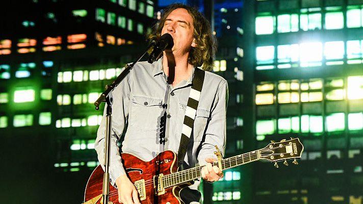 The photo shows a musician performing live on stage at night. He’s playing a reddish‑brown electric guitar with white accents and singing into a microphone. He’s wearing a light blue button‑up shirt, and his shoulder‑length hair falls naturally as he leans into the performance. Behind him, tall buildings with illuminated windows create a vibrant cityscape backdrop