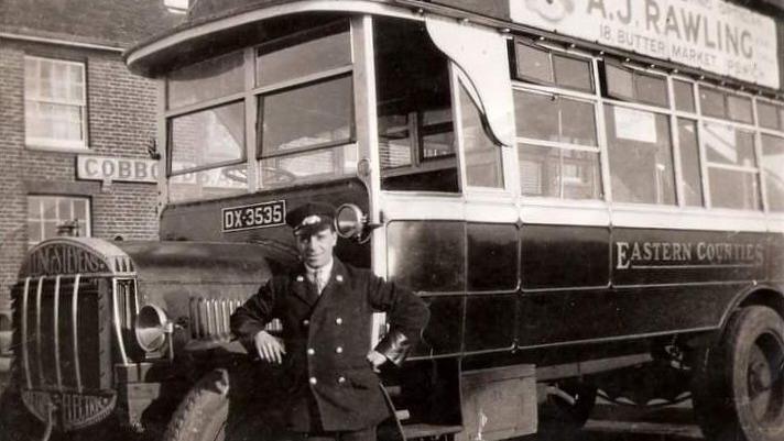 A black and white photo from 1922. It shows a bus driver standing in front of the bus. He is stood by the engine and wearing a bus driver's uniform with a hat.