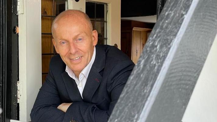David Burns wears a white shirt and black suit. He looks through the door of his home in Kempston.