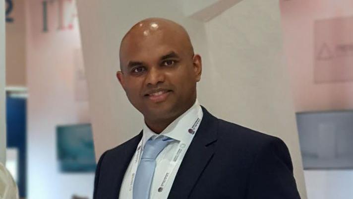 Dr Vivek Koncherry smiles wearing a tie and a navy suite in an academic building. 