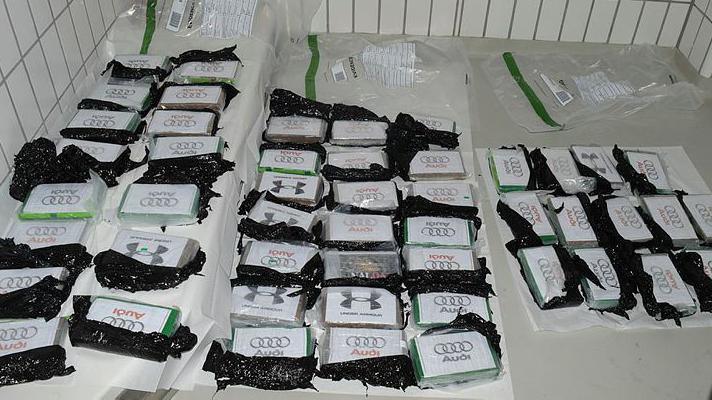 A white table or surface in a white tiled room, with a series of small plastic packets torn open with the AUDI logo on lined up neatly. Presumably containing cocaine/drugs. Three empty plastic evidence bags lie flat in the background.