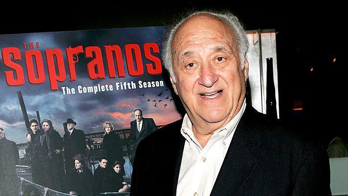 Jerry Adler, an elderly man, pictured at an evening event for The Sopranos TV series. He has short grey hair and wears a collared white shirt under a black jacket. He is stood in front of a poster for The Sopranos which reads: "The Sopranos: The complete fifth season"