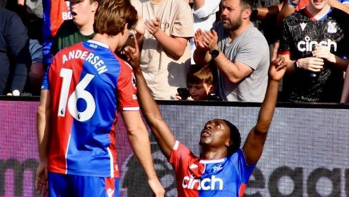 A photo of a Crystal Palace football player kneeling on the pitch in celebration with arms raised, while another teammate stands nearby. Both players are wearing Crystal Palace's red and blue kit. In the background, spectators are cheering in the stands.