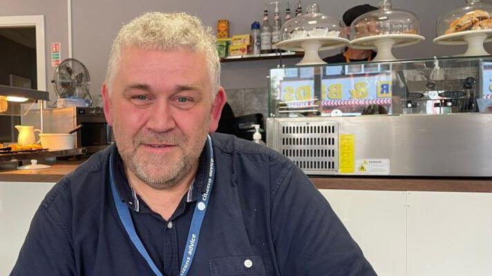 A head and shoulders photo of Dave Pearson who is smiling at the camera. He has short blond curly hair and is wearing a navy-blue shirt and a blue lanyard. He is sitting in a cafe, in front of the counter. Kitchen equipment and cake stands can be seen in the background. 