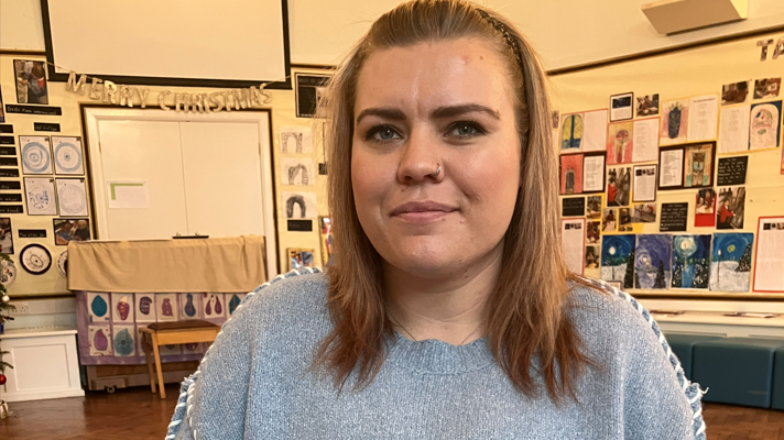 A head and shoulders shot of Charlotte Hornsby who is a young woman wearing a blue jumper sitting in a classroom with pictures by children on the walls 