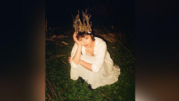 A woman is crouching in the dark on some grass. She has short, dark hair and is wearing a white dress and a crown on her head.