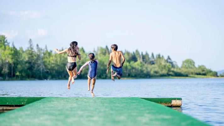 A small group of three children are seen in mid air jumping off the end of a dock on a sunny summer day. They are each striking a fun pose before they hit the waters surface to make a splash