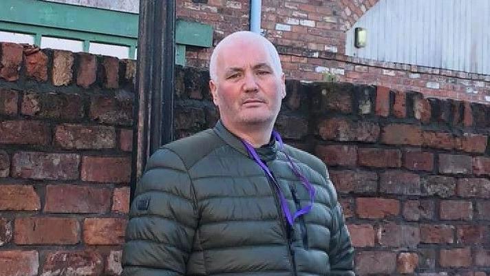 Gary Nicol leaning against a brick wall wearing a green jacket and purple lanyard.