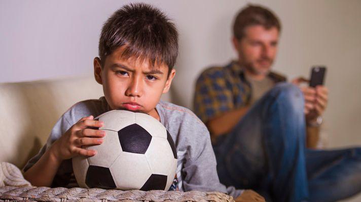 Boy waiting to play football with his dad, who's on his phone.