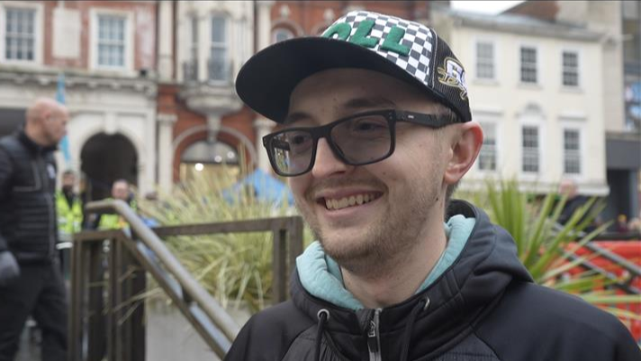 Sam Broom looks to the left as he smiles. He wears a cap, glasses which are slightly tinted and a black coat.