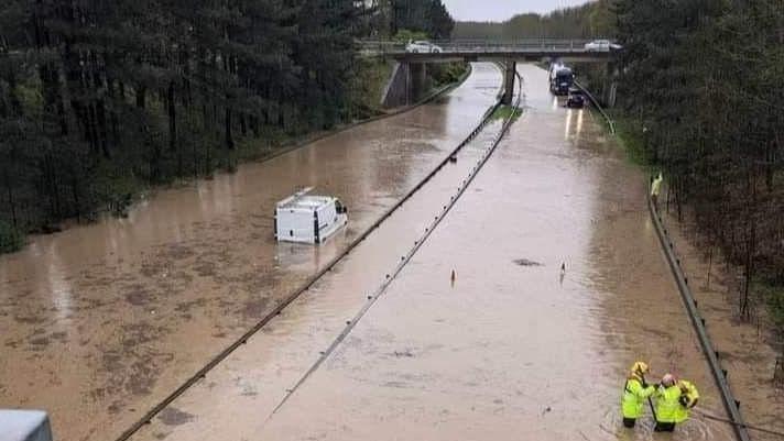 The image shows the A189 about two feet deep in water with a van submerged and two men wearing yellow jackets.