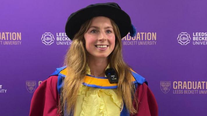 A woman in a cap and gown stands in front of a purple background as she receives her honorary doctorate.