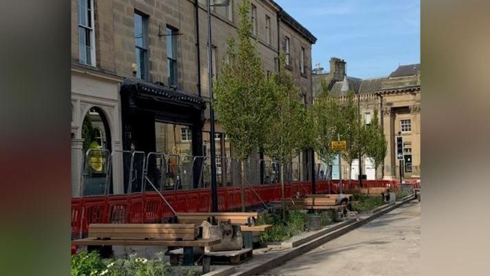 Wooden benches with the new trees and plants. Orange fencing still surrounds the area