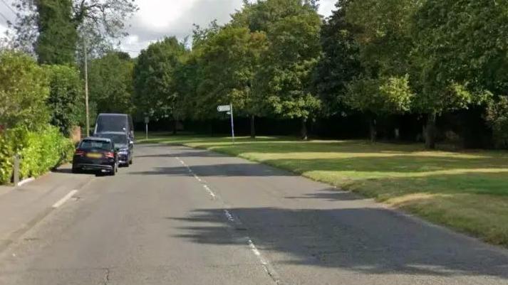 A quiet road, bordered by hedges on one side and a grassy bank and trees on the other. Three vehicles are parked on the kerb.