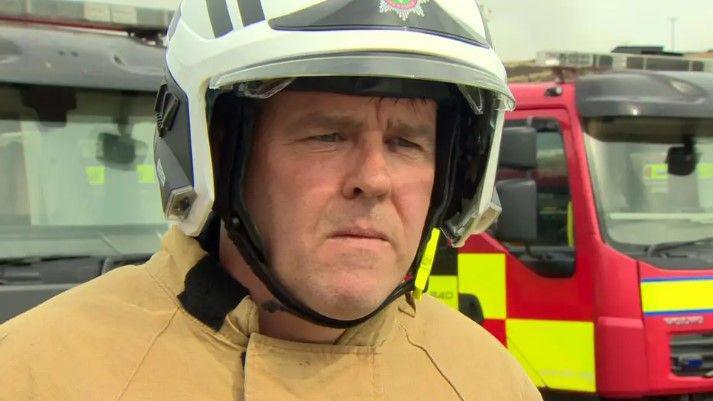 A firefighter is wearing a white helmet and a heavy beige jacket. Fire engines are parked behind him.