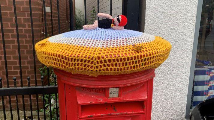 Postbox topper showing a red post box with a knitted doll in a swimming position. The rest of the topper is blue, white and gold. 