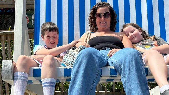 Gemma and her kids sitting on a giant deck chair. Gemma sits in the middle and is wearing a black top, blue jeans and sunglasses. Her daughter sits on her left side, while her son sits on her right.