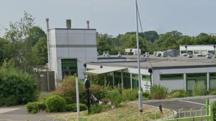 A google maps image of a white and green school building and partial carpark beyond a green fence