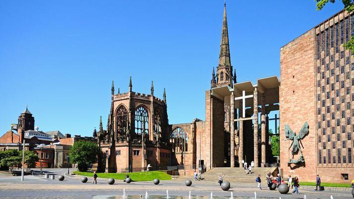 Coventry's old cathedral ruins seen next to the new 20th Century building. There is a clear, blue sky and people are walking around the cathedral precincts. There are decorative globes and a water feature is visible.