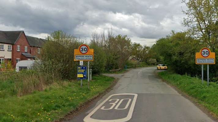 A country lane, with a large white 30 sign painted on the road, and yellow signs either side, each with a 30 sign, and the words Bomere Heath, please drive carefully.

Modern terraced housing can be seen to the left of the road