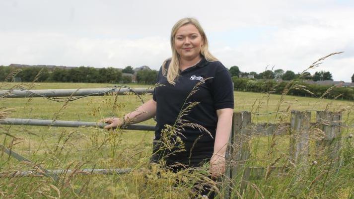 Emily Routledge is standing smiling in a field with long grass. Her hand is on a metal fence and she is wearing a DWT black t-shirt.